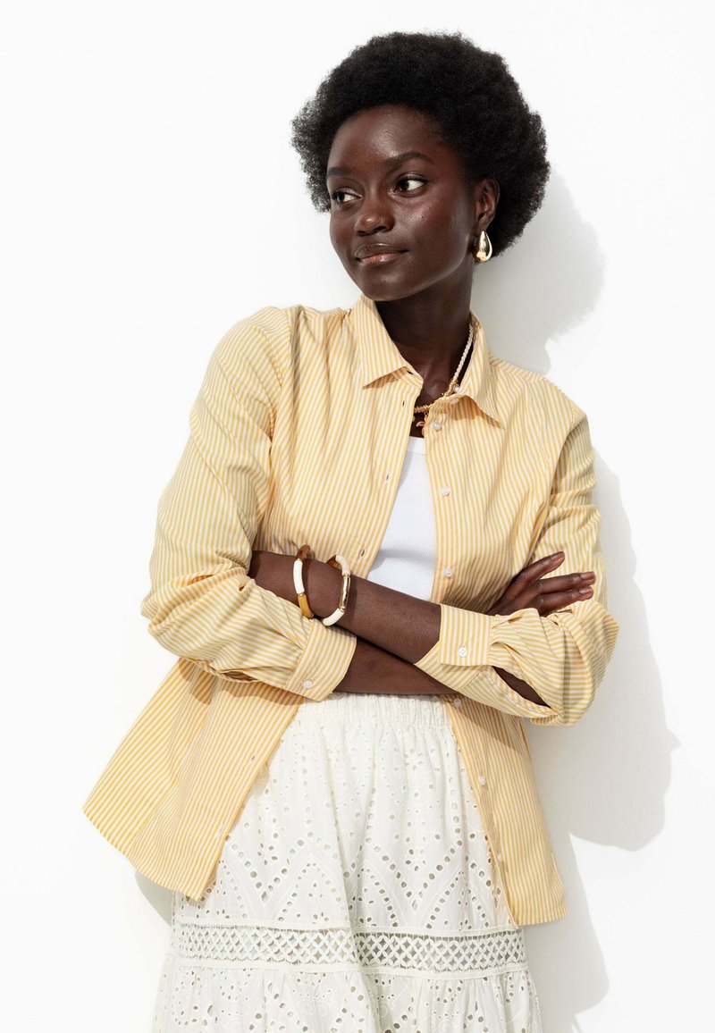 Chemise boutonnée à rayures jaunes et blanches avec des manches longues, portée par-dessus un débardeur blanc et une jupe blanche texturée avec des détails en œillets.