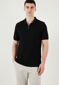 Čierna polokošeľa s golierom na zips, krátkymi rukávmi a hladkou textúrou. Nosená s svetlými nohavicami. Jednoduchý a elegantný dizajn.