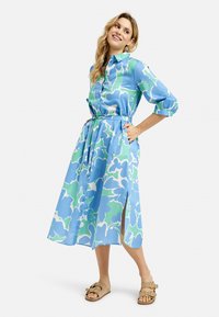 Blau- und grün gemustertes Kleid mit floralem Druck, geraffter Taille, Knopfleiste und seitlichen Schlitzen; kombiniert mit braunen Sandalen.