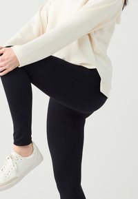 Leggings noirs à la coupe ajustée et texture lisse, accompagnés d'un sweat-shirt ample de couleur crème et de baskets blanches.