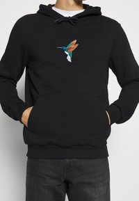 Schwarzer Hoodie mit Kängurutasche, ausgestattet mit einem bunten gestickten Kolibri auf der Brust, aus weichem Stoff gefertigt und mit gerippten Bündchen.