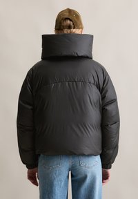 Melns pūkains jaka ar augstu apkakli, ar gludu tekstūru un elastīgiem rokturiem, uzvilkta virs gaiši zilām džinsu biksēm.