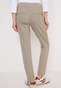 Person in beigen, schmal geschnittenen Hosen mit Gesäßtaschen und weißen Sneakern mit burgunderroten Akzenten, stehend vor einem schlichten Hintergrund.