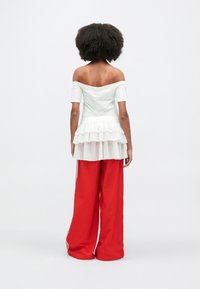 Λευκό τοπ με αποκαλυπτικούς ώμους και στρώση από διαφανές βολάν στο τελείωμα, συνδυασμένο με φαρδιές κόκκινες παντελόνες με λευκές ρίγες στο πλάι.