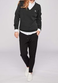 Pull en maille vert foncé avec col en V, chemise blanche à col en dessous, associé à un pantalon de jogging noir et des baskets blanches.