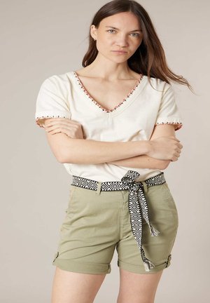 Femme aux longs cheveux bruns portant un chemisier crème à col en V avec des bords cousus, un short vert olive et une ceinture en tissu noir et blanc à motifs.