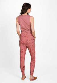 Woman standing, wearing sleeveless pink striped top and pink floral patterned pants, facing away with hands in pockets.