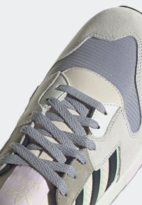 Ljust grå sneaker med en texturerad suede- och mesh-övre del, med grå snören, gröna accenter och en kontrasterande svart randdesign.