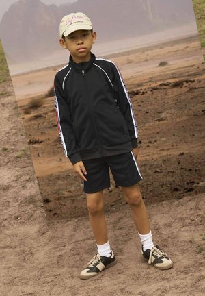 Giacca nera con zip e strisce bianche e rosse, abbinata a pantaloni corti neri. Indossa un cappellino beige e sneakers bicolori. Il terreno è asciutto e sabbioso.