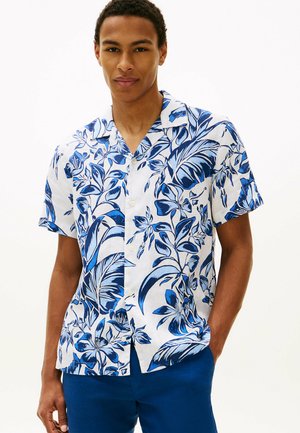 Młody mężczyzna ubrany w białą koszulę z krótkim rękawem ozdobioną niebieskimi wzorami kwiatowymi oraz niebieskie spodnie, stoi z jedną ręką w kieszeni.