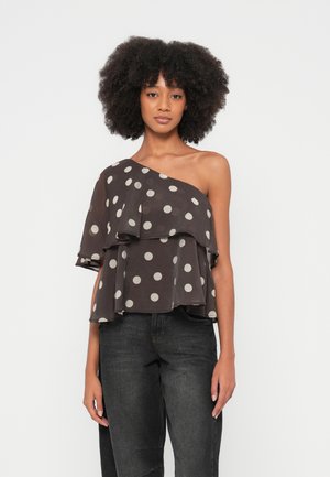 Kvinde med krøllet hår iført sort one-shoulder top med hvide polkaprikker og mørke jeans, stående foran en ensfarvet lys baggrund.