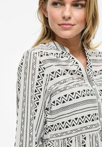 Blouse à motifs noir et blanc avec des imprimés géométriques, un décolleté en V et une fermeture à boutons, confectionnée dans un tissu léger et texturé.