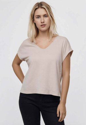 Mujer rubia con ojos azules lleva una camiseta beige de manga corta con cuello en V y pantalones negros, de pie con una mano en la cadera.