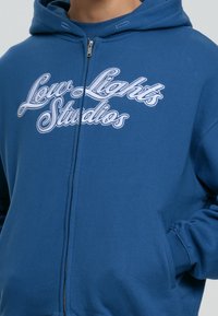 Blauwe ritshoodie van zacht materiaal, met op de borst in witte geborduurde tekst "Low Lights Studios", uitgerust met een kangoeroezak en een capuchon.