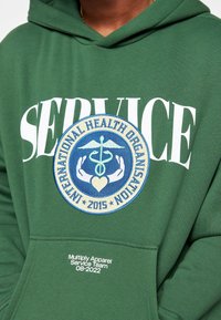Felpa verde con testo bianco "SERVICE," un patch circolare ricamato con il simbolo del caduceo e "ORGANIZZAZIONE MONDIALE DELLA SANITÀ."