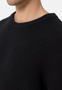 Svart ribbad tröja med rundad halsringning, med ett texturerat stickat mönster och synlig söm längs halslinningen.