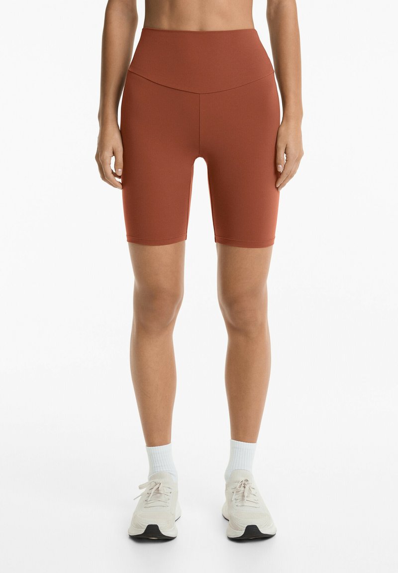 Burgundy cykelshorts med hög midja och sömlös design, slät textur och längd ovanför knäet, som kombineras med ljusa sneakers och ankelsockor.