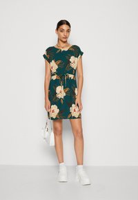 Vestido floral em azul-petróleo com mangas curtas, cintura ajustada e cordão. Apresenta padrões de flores em bege e dourado. Usado com ténis brancos.