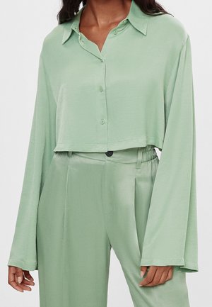 Femme portant une chemise ample courte à manches longues de couleur vert clair avec des boutons et un pantalon taille haute assorti.
