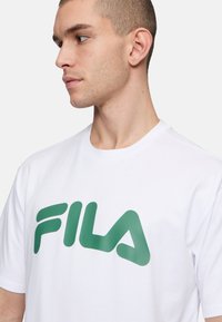 Camiseta de algodón blanca con un gran logo verde de FILA en el frente, mangas cortas, cuello redondo y textura suave.