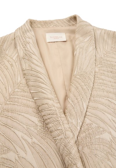 Blazer beige testurizzato con motivi a linee astratte in rilievo e etichetta "SLOWEAR Montedoro" all'interno del colletto.