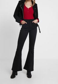Jean noir évasés et un haut rouge à manches longues côtelé sous une veste en cuir noire avec des détails en métal argenté et une silhouette ajustée.