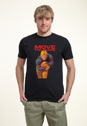 Homme blond portant un T-shirt noir avec un joueur de football vintage tenant un ballon et le texte rouge « MOVE THE CHAINS », les mains dans les poches d'un pantalon kaki.