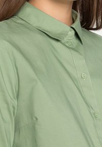 Ljust grön skjorta med krage i ett mjukt tyg, med en knapplist framtill och subtila sömnadsdetaljer nära knapparna.