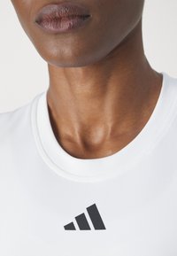 Valkoinen muotoiltu urheilupaita, jossa on pyöreä pääntie ja musta Adidas-logo rinnassa, valmistettu sileästä ja joustavasta kankaasta.