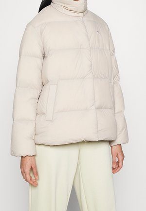Femme portant une doudoune matelassée beige clair avec un col montant et des poches latérales, associée à un pantalon de couleur crème.