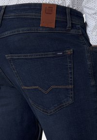 Dunkelblaue Jeans aus Denim mit kontrastierenden Nähten, einer Gesäßtasche, einem Lederetikett und einem weißem Hemd, das eingesteckt ist.