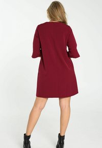 Femme aux cheveux blonds portant une robe bordeaux longueur genoux et des bottines noires, debout dos à la caméra.