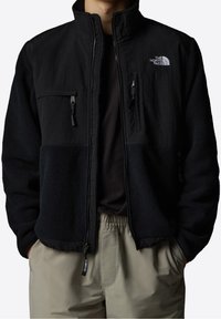 Czarna polarowa kurtka z gładkim materiałem na górze, kieszeniami na suwak i wysokim kołnierzem; na lewej piersi logo The North Face.