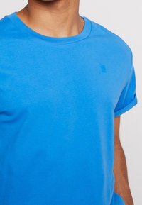 Blå bomullströja med rund halsringning och uppvikta korta ärmar. Har en diskret logotyp på bröstet i matchande blå färg.