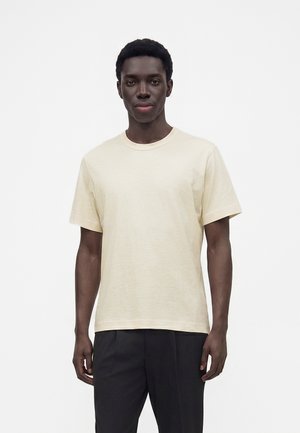 Ung mand med kort krøllet hår iført en cremefarvet kortærmet t-shirt og sorte bukser, stående foran en ensfarvet hvid baggrund.