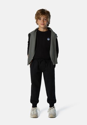 Felpa nera, hoodie senza maniche grigia, pantaloni jogger neri e sneakers chiare. Outfit casual con un piccolo logo rotondo sulla felpa.
