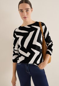 Schwarzer und weißer Pullover mit Zickzackmuster, runder Halslinie und gerippter Struktur, kombiniert mit dunkelblauen Jeans und einer braunen Tasche.