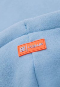Ljusblå sweatshirt tyg med en orange rektangulär etikett som visar "LEVEL UP" i vit text och en logotyp i övre vänstra hörnet.