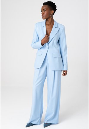 Kobieta w jasnoniebieskim, dopasowanym garniturze z szerokimi spodniami i marynarką, ozdobiona dużymi kolczykami-kołami, patrzy w lewo.