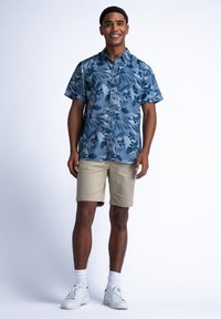Camisa azul de botones de manga corta con un patrón de hojas, combinada con pantalones cortos beige, calcetines blancos y zapatillas blancas.