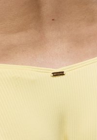 Κοντινό πλάνο του λαιμού και του άνω μέρους του στήθους ενός ατόμου που φοράει κίτρινη ριμπ μπλούζα με μια μικρή χρυσή ετικέτα "Hollister" στο λαιμό.