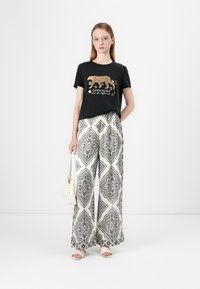T-shirt noir avec imprimé léopard doré et texte, associé à un pantalon large blanc featuring un motif géométrique noir. Sac beige et sandales.