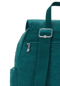 Mochila verde azulado con correa ajustable, hebilla plateada, bolsillo frontal con cremallera y pequeño compartimento lateral sobre un fondo blanco.