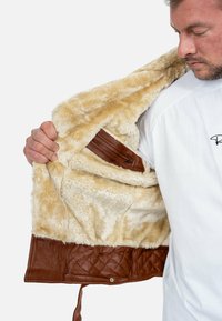 Giubbotto in pelle marrone con fodera in pelliccia sintetica beige, dotato di una tasca con zip e texture trapuntata sull'orlo. Aperto per mostrare i dettagli interni.