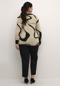 Pull en tricot beige avec motif abstrait noir, col rond et poignets côtelés. Associé à un pantalon noir et des talons noirs.