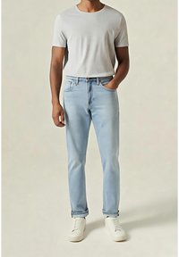 Homme portant un t-shirt gris clair à manches courtes, un jean bleu clair avec revers et des baskets blanches, debout avec une main dans la poche.