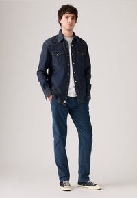 Bărbat în picioare cu mâinile în buzunare, purtând o cămașă din denim închis, un tricou alb, pantaloni blue și adidași negri, pe un fundal simplu.