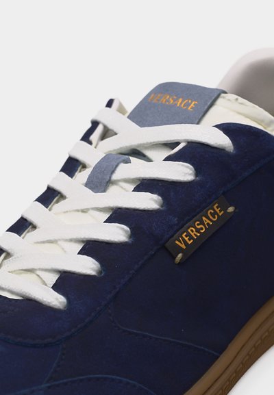 Marineblaue Versace-Sneaker mit weißen Schnürsenkeln, hellbrauter Sohle und Versace-Logo in Gold auf der Zunge und der Seitenfläche.