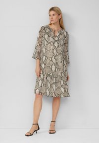 Beige und braunes Schlangenmuster-Kleid, leichtes Material, gestuftes Rockdesign, runder Ausschnitt mit Bindebändern, Dreiviertelärmel, getragen mit schwarzen High Heels.