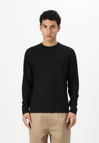 Black long-sleeve shirt featuring a textured pattern on one side, round neck design, made of soft fabric, paired with beige trousers.